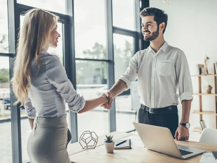 Smiling man and woman shaking hands at a desk in a modern office, symbolizing partnership, featured on towndirectorynet.com